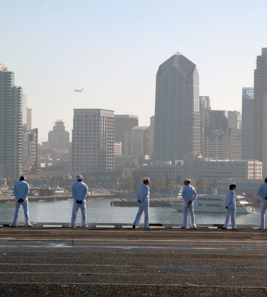 Sailors man the rails of an aircraft carrier with the downtown San Diego skyline behind them